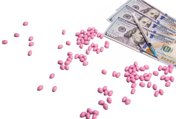 Scattered pink pills on a white background next to $300 US dollar bills and a syringe. The importance of being healthy to save money on medicine. High insurance costs. Prescription medicine bills. 