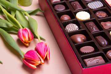 Perfect gift for special occasions, a red box of assorted chocolate on a table next to pink tulips.