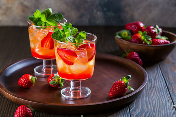 Red cocktail with ice, mint leaves and strawberry on wooden background.