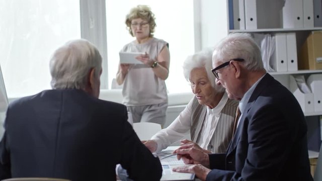 Medium shot of team of elderly businesspeople in formalwear sitting at desk and discussing documents in office; senior woman in glasses working on tablet beside window