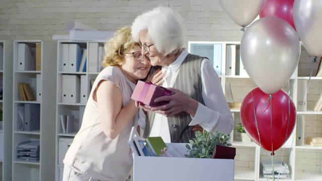 Medium shot of happy mature businesswoman congratulating retiring female colleague packing things in office: she giving her present and hugging