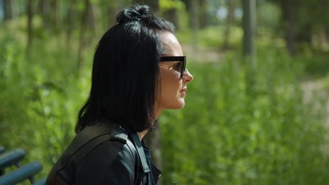A profile of a black haired woman wearing sunglasses, sitting down on a bench in the park.