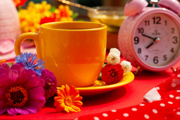 Breakfast with tea is surrounded by bright colors. A cup of tea, a clock, a silk scarf, flowers for rest in spring and summer. Morning tea in colorful tones.