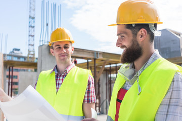 Two young construction workers wearing yellow hard hats and reflective safety vests while analyzing together the plan of a new building