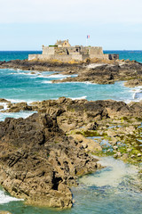Fototapeta na wymiar The Fort National built by french military architect Vauban on a tidal island, seen from the city of Saint-Malo, France, with the french flag blowing in the wind and rocks in the sea in the foreground