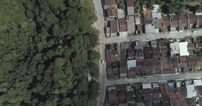 Aerial zenital drone shot over a car. Road centered, forest in one side, humble community in another side.