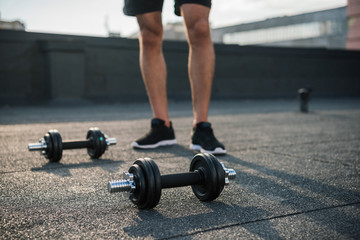 cropped image of sportsman standing near dumbbells on roof