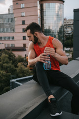 handsome sportsman sitting on roof railing with bottle of water and looking down