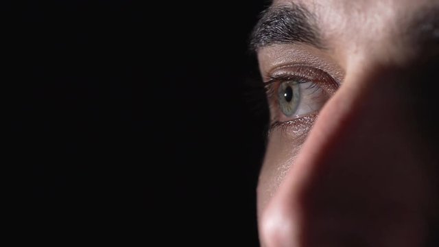 Portrait extreme closeup of european man 20s moving green eyes and blinking while looking aside in fear, isolated over dark background
