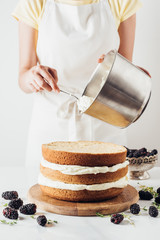 cropped shot of woman pouring cream onto freshly baked cake on white