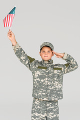 portrait of boy in camouflage clothing saluting while holding american flagpole in hand isolated on grey