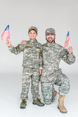 smiling father and son in camouflage clothing with american flagpoles in hands on grey background