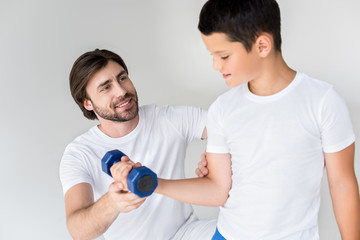 father helping little son with dumbbell to exercise on grey background