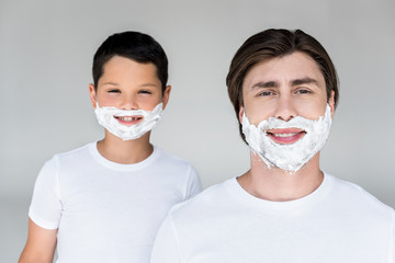 portrait of smiling father and son with shaving foam on faces isolated on grey