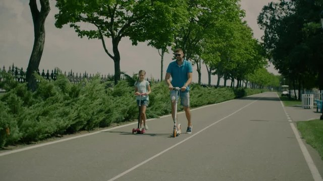 Positive handsome father in sunglasses and cute girl riding kick scooters together in summer park. Active family enjoying leisure, riding scooters while spending great time together outdoors. Slo mo.