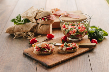 Delicious slices of cut bread covered with chopped cream cheese, cherry tomato and prosciutto and whole sandwiches in the background wrapped in paper bag.