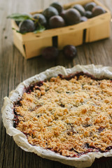 Delicious homemade plum pie with crumbles on top and whole plum fruit in the wooden basket in the background.