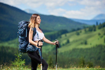 Close-up portrait of beautiful sporty woman traveller with blue backpack and trekking sticks, hiking on the top of a hill, looking away, enjoying sunny day in the mountains. Copy space