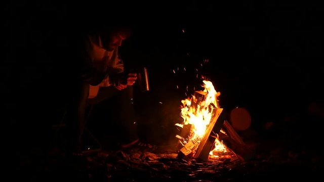 bearded men drink tea from big cup and make a photo on a cellphone of bonfire in a dark. slow motion