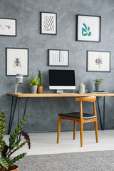 Gallery of posters above desk with computer desktop in grey home office interior with chair. Real photo