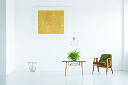 Lamp above table with fern next to green armchair in white interior with yellow painting. Real photo