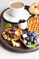 Belgian waffles with fresh blueberries, peaches, chocolate sauce and a cup of coffee on a rustic wooden cutting board. Liege style belgian waffles. Breakfast or lunch concept.