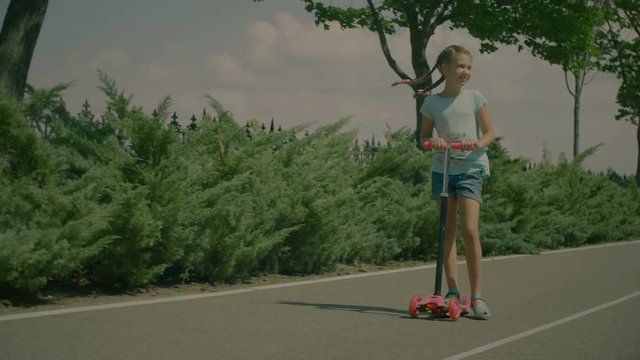 Happy little cute girl learning to ride and balance on push three wheel adjustable height scooter in summer park over beautiful landscape background. Excited child riding kick scooter outdoors. Slo mo
