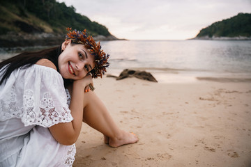 A girl in a white sarafan and with a wreath on her head smiles and looks romantic, sitting on the sand, against a background of tropical landscape, sea, and sunset.