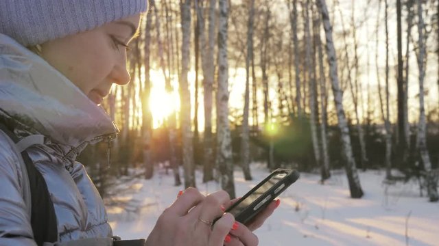 Woman is walking in the woods. Traveler is photographed on the phone in forest. Girl does selfie and communicates with her smartphone. Evening time with beautiful juicy sunset. Explorer is dressed in