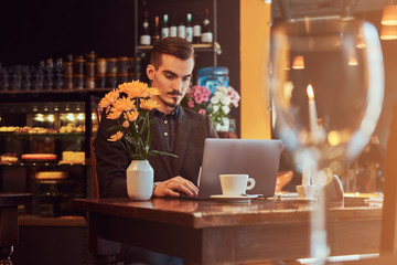 Handsome freelancer man with stylish beard and hair dressed in a black suit working on laptop while sitting at a cafe.