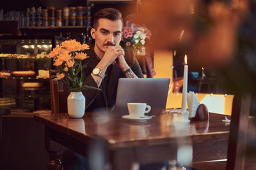 Portrait of a pensive handsome businessman with stylish beard and hair dressed in a black suit sitting with hand on chin at a cafe with an open laptop, lookong at the camera.