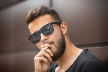 A young handsome white bearded man in sunglasses and black t-shirt smokes a cigarette in the street in the spring. Close up.