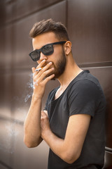 A young handsome white bearded man in sunglasses and black t-shirt smokes a cigarette in the street in the spring. Close up.