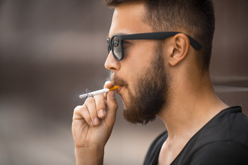 A young handsome white bearded man in sunglasses and black t-shirt smokes a cigarette in the street in the spring. Close up.