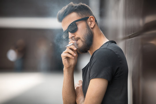A young handsome white bearded man in sunglasses and black t-shirt smokes a cigarette in the street in the spring. Close up.