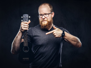 Redhead bearded male musician wearing glasses dressed in a gray t-shirt holds ukulele. Isolated on a dark textured background.