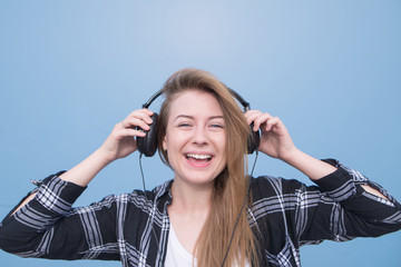 Close portrait of a happy girl who listens to music in headphones on a blue background, looks into the camera and smiles. Young woman listens to music in the headphones isolated on a blue background