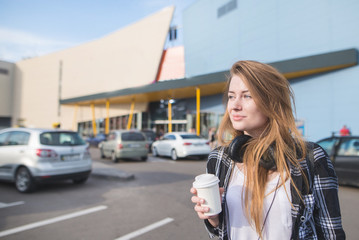 Attractive girl stands with a cup of coffee in the hands of the background of modern architecture and looks out of the way. Pretty girl with coffee in her hands walking around the city
