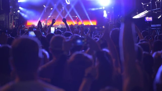 Slow motion shot of unidentified rock band performing in front of excited fans crowd who raising hands, applauding and shooting mobile videos