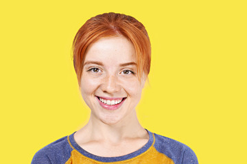 Beautiful redheaded young woman with satisfied facial expression, isolated on yellow background. Attractive female with natural red hair & brown eyes showing sincere smile & perfect teeth. Copy space.