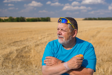 Outdoor portrait of a bearded senior in sunglasses taking apricots in the hand against blue cloudy sky and agricultural field