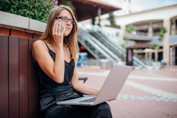 Young woman sitting on bench in park on summer day and reading text message on cell phone while using silver laptop outdoors in the city space