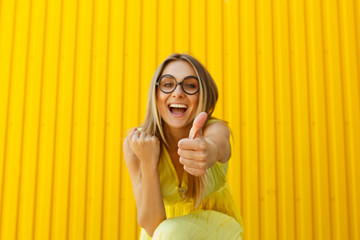 Portrait of a joyful girl wearing toy funny glasses looking up over yellow background showing hand ok sign