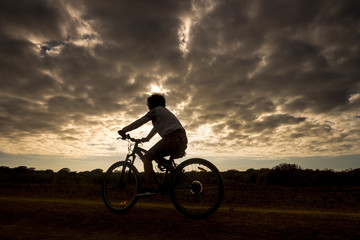 mountain biker woman in action with golden beautiful sunset in background, silhouette of athlete going on bike following the road and enjoying the summer time
