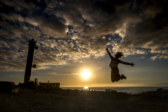 happiness concept for middle age woman jumping with beautiful dark and colored sunset and lighthouse in background. vacation and winner time for happy people