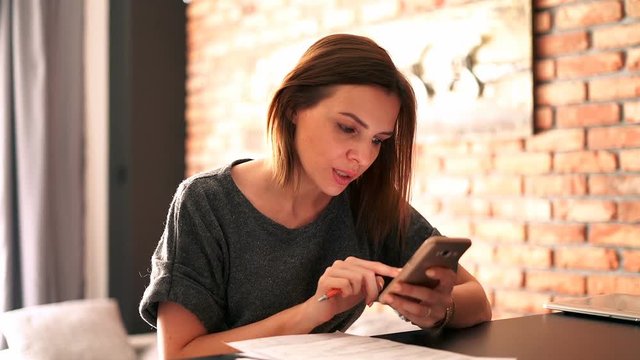 Businesswoman comparing documents with smartphone by table at home
