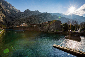 Sunny winter day cityscape of the old Kotor town with the North defensive walls and the crystal green waters of Scurda river. View with the Sun rays and light reflections