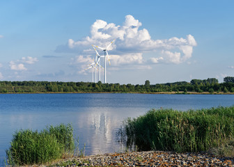Several wind turbines stand in front of a huge cloud formation at the shore of a lake, warm summer...