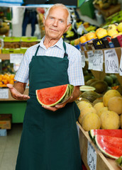 Positive senior male owner of greengrocery shop in apron offering fresh fruits and vegetables