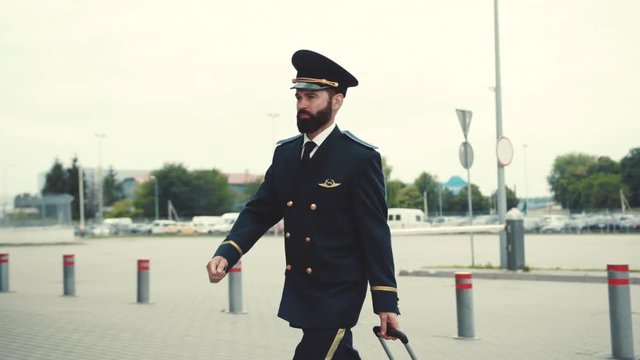 Handsome caucasian bearded pilot puts on a cap and pulls his luggage. Travel status, profession concept. Business trip, being on assignment. Male portrait